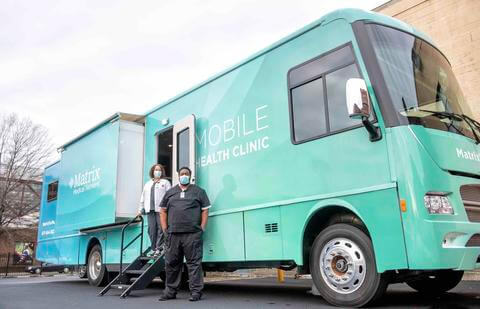 A mobile health clinic truck with two health workers standing near it.