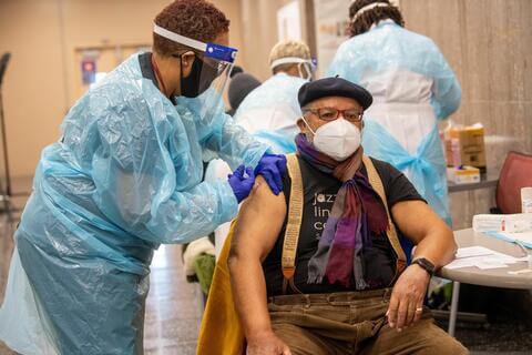 A nurse in personal protective equipment injecting an older patient with a COVID-19 vaccine.