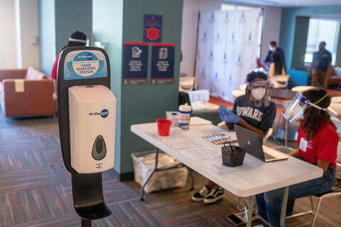 The interior of a Howard University dormitory, with a hand sanitizer station in the foreground and students in PPE in the background.