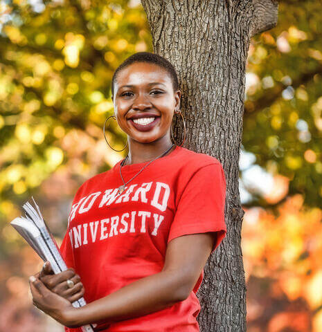 A young woman in a Howard University t-shirt leaning against a tree and smiling as she holds books.