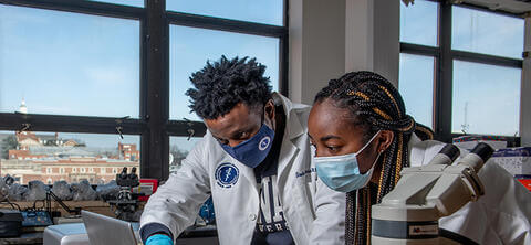 Howard students in labcoats seated in a classroom using a computer and microscope to evaluate medical data.