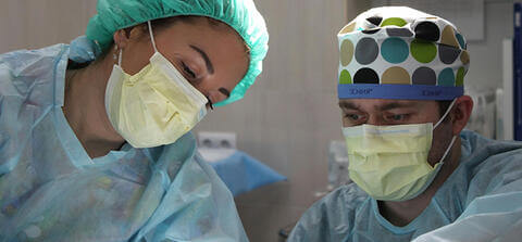 Two medical professionals in scrubs and wearing masks work together in a lab, leaning in to look at what they're studying.